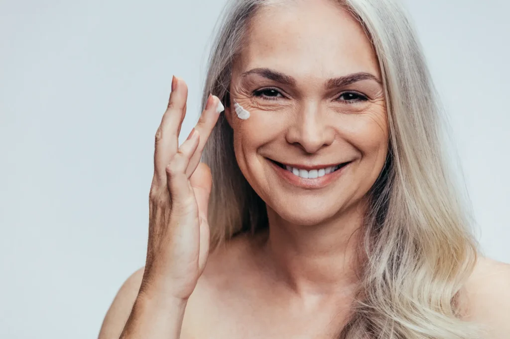 Mature woman smiling while applying cream to cheek with fingers