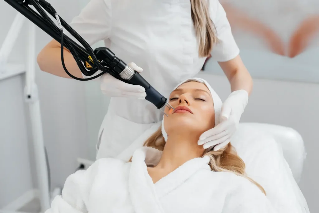 Gloved practitioner holds a laser device near a woman's upper lip as she lies on a treatment chair.