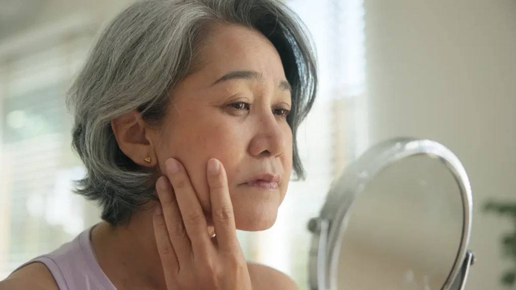 Mature woman touches her cheek while examining her face in a tabletop mirror near a window.