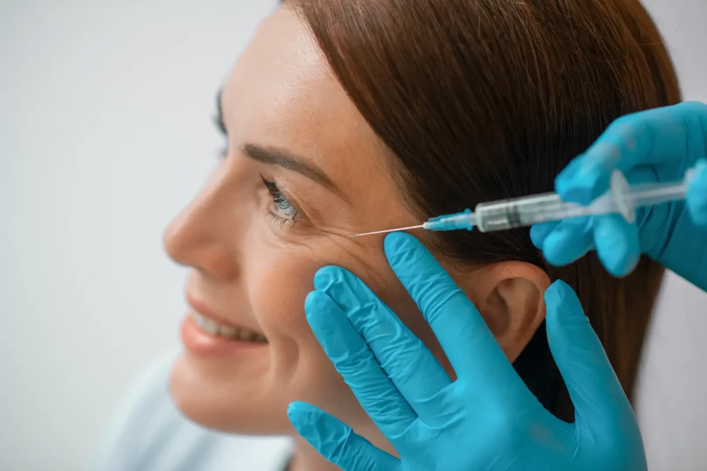 Gloved practitioner positions a syringe near the outer corner of a woman's eye during a facial procedure.