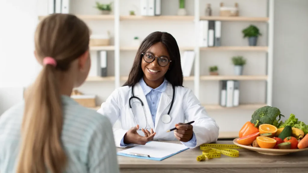 Doctor speaks with a patient across a desk with fresh produce, a measuring tape, and paperwork nearby.