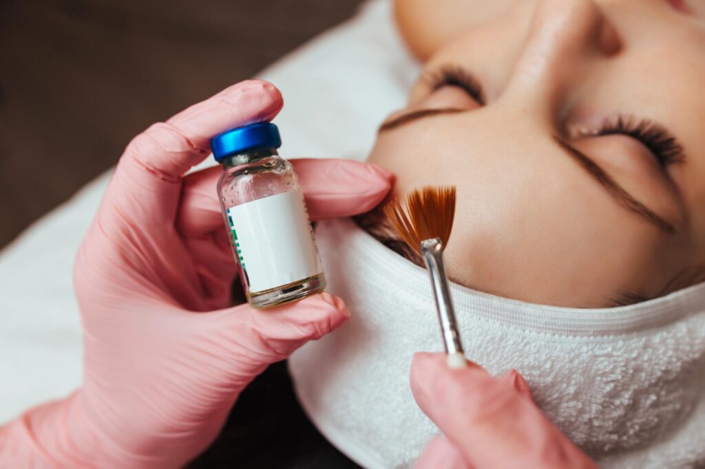 Gloved hands hold a small vial and fan brush beside a woman’s cheek during a facial treatment.