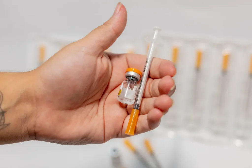 Hand holds a small medication vial and syringe with orange accents against a blurred clinical background.