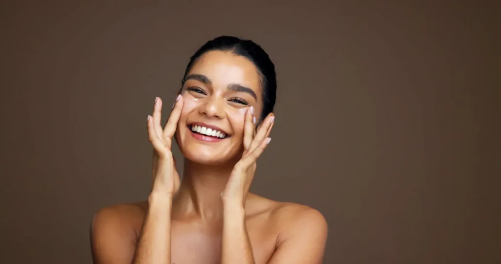 Smiling woman applies a white skincare product beneath both eyes with her fingertips.